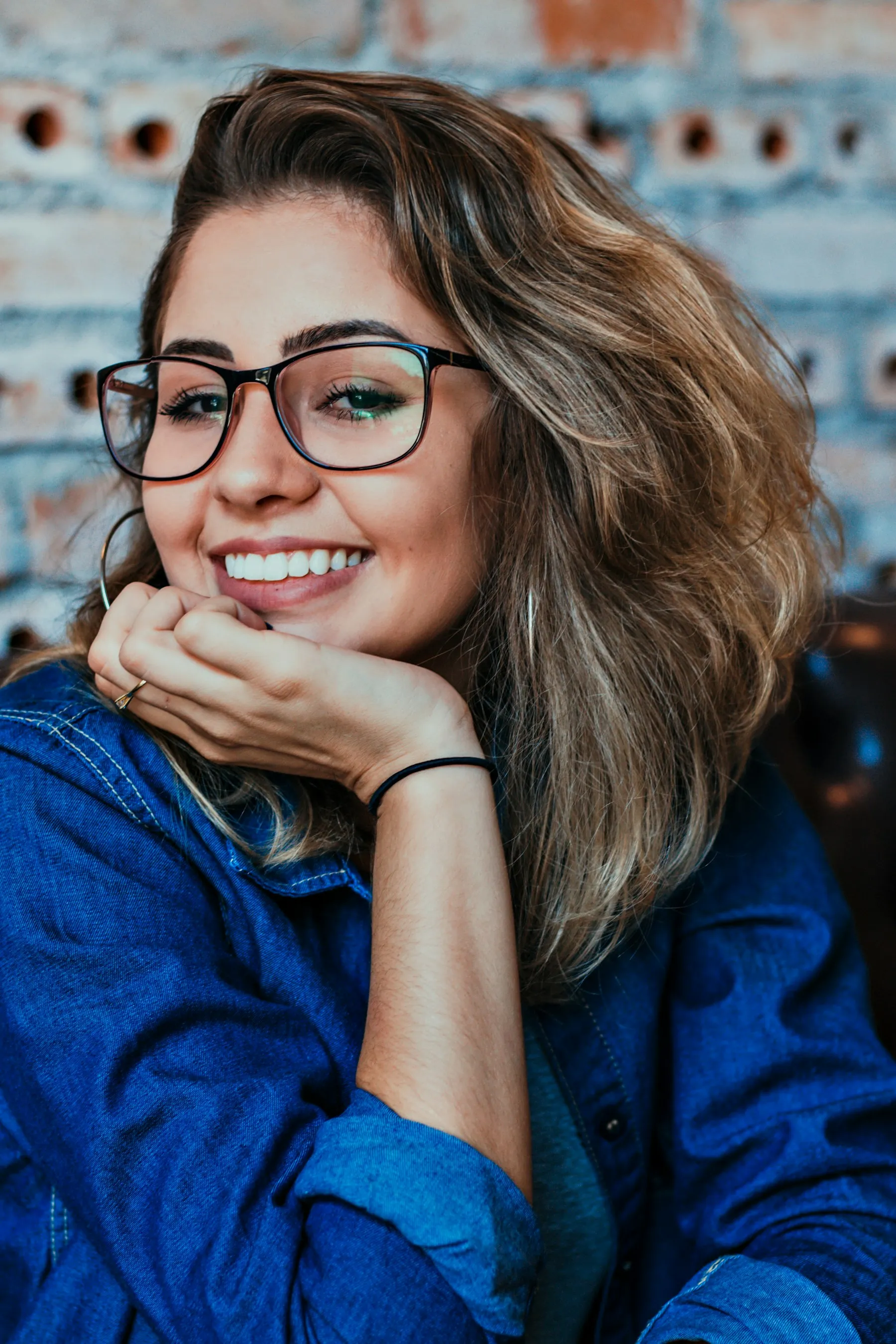 Mulher sorrindo de óculos de armação escura, com a mão no queixo