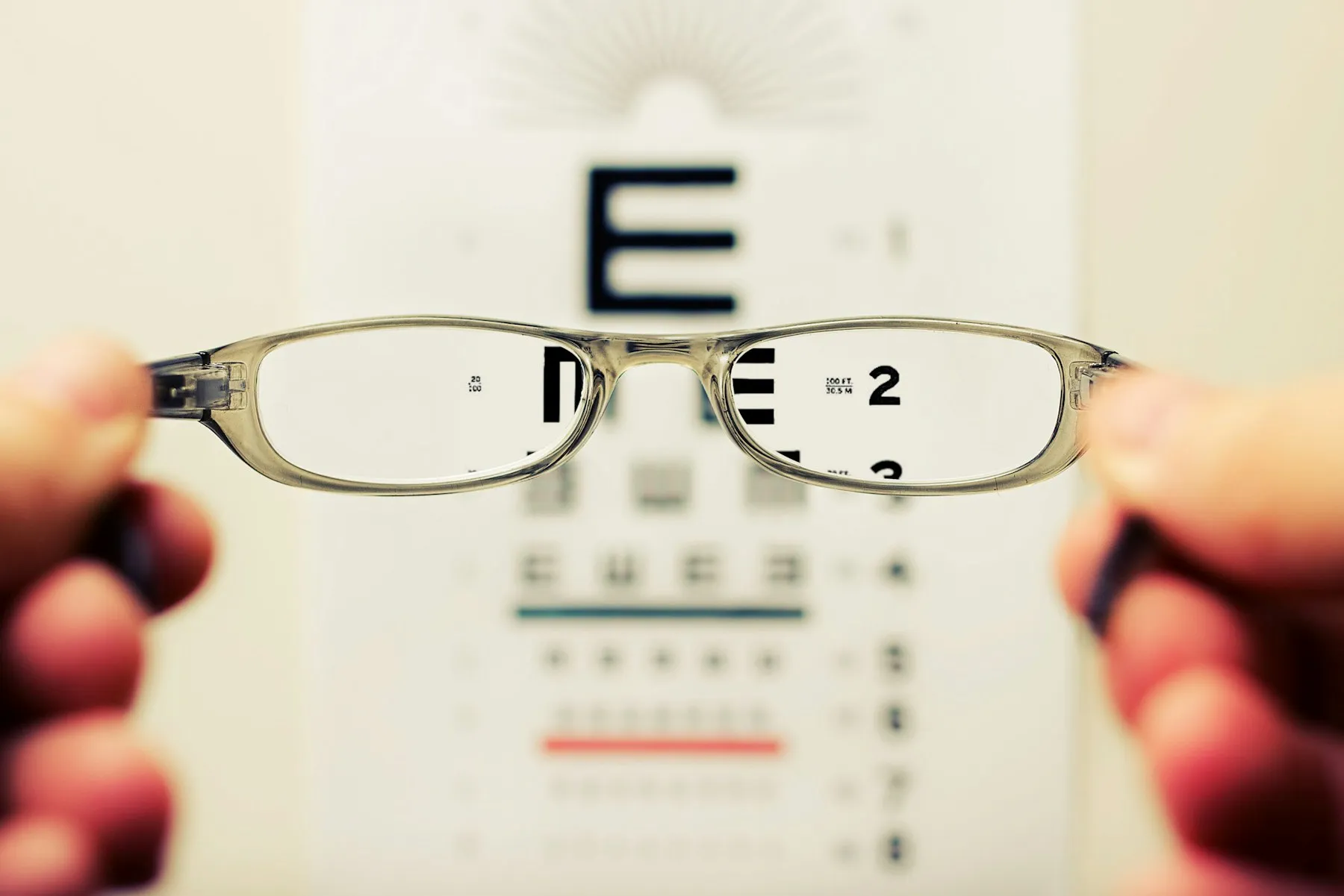 Mãos segurando um par de óculos diante de uma tabela de letras, que aparece nítida através das lentes